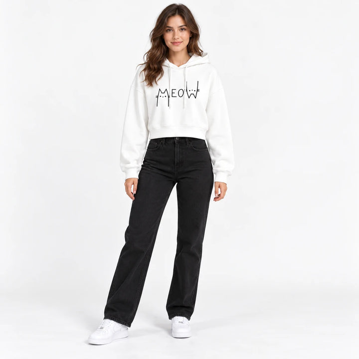 White Meow graphic cropped hoodie on a full-body model with long sleeves and a drawstring hood with black jeans and sneakers.
