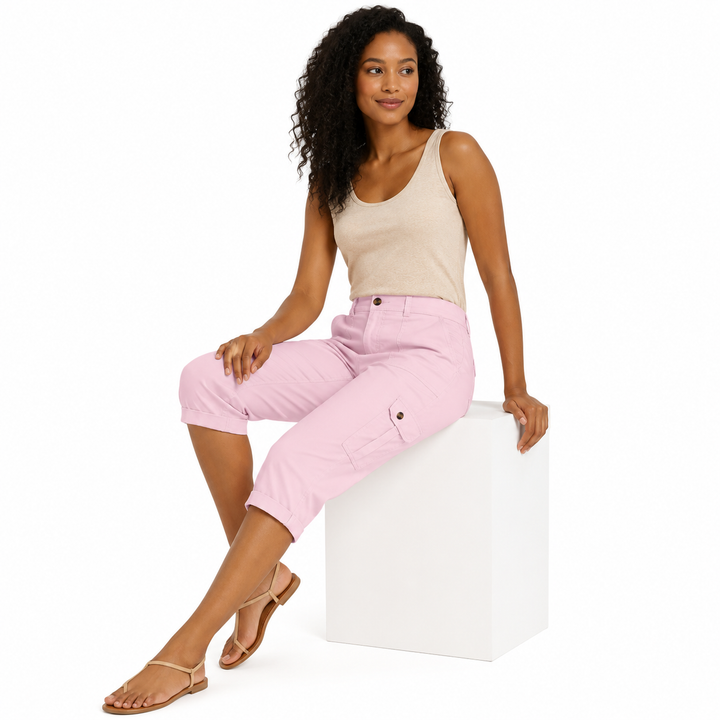 A woman with curly dark hair sits on a white cube against a white studio background, wearing a light beige tank top, light pink cropped cargo pants, and tan flat sandals.