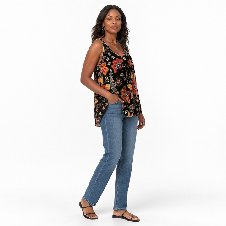 A woman modeling a sleeveless black tank top with a colorful floral and butterfly print, paired with blue jeans and black sandals, against a plain white studio background.