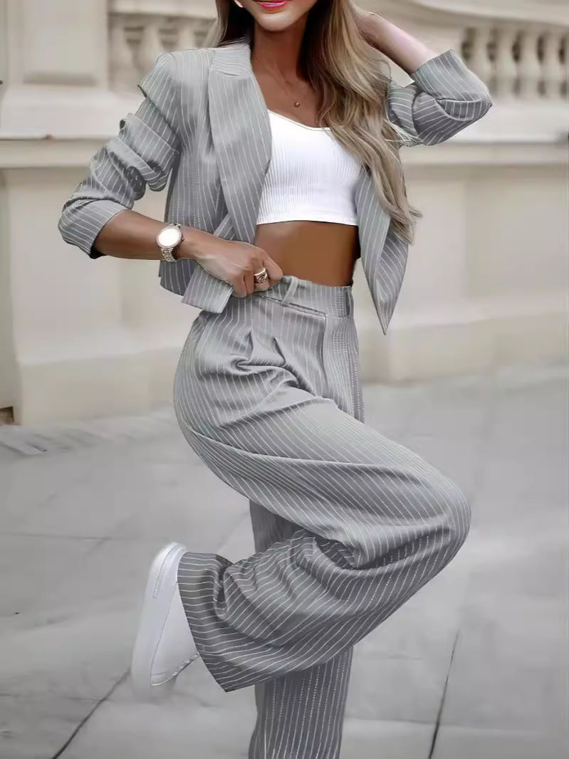 Gray pinstripe cropped jacket and pants shown on an outdoor model in a bent-leg pose, with a white inner top and sneakers.