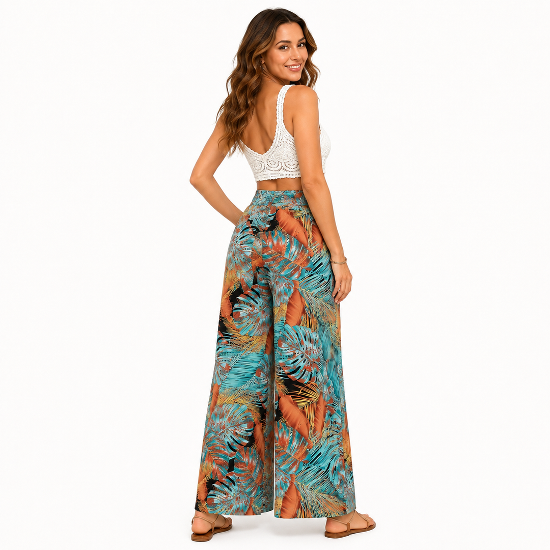 A woman stands with her back to the camera wearing high-waisted wide-leg pants with a vibrant tropical leaf print in teal, orange, and brown, paired with a white crochet crop top and brown flat sandals, on a solid white background.