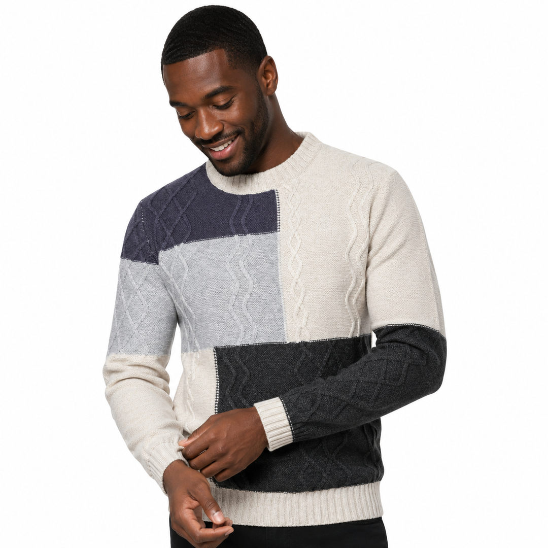 A man wearing a color-blocked cable-knit sweater with cream, light gray, navy, and charcoal sections, shown against a plain white background.