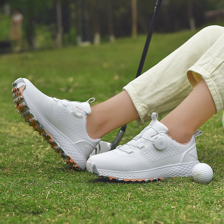 White and pink rotary buckle shoes worn while seated on grass beside a golf ball, showing the side profile and patterned sole