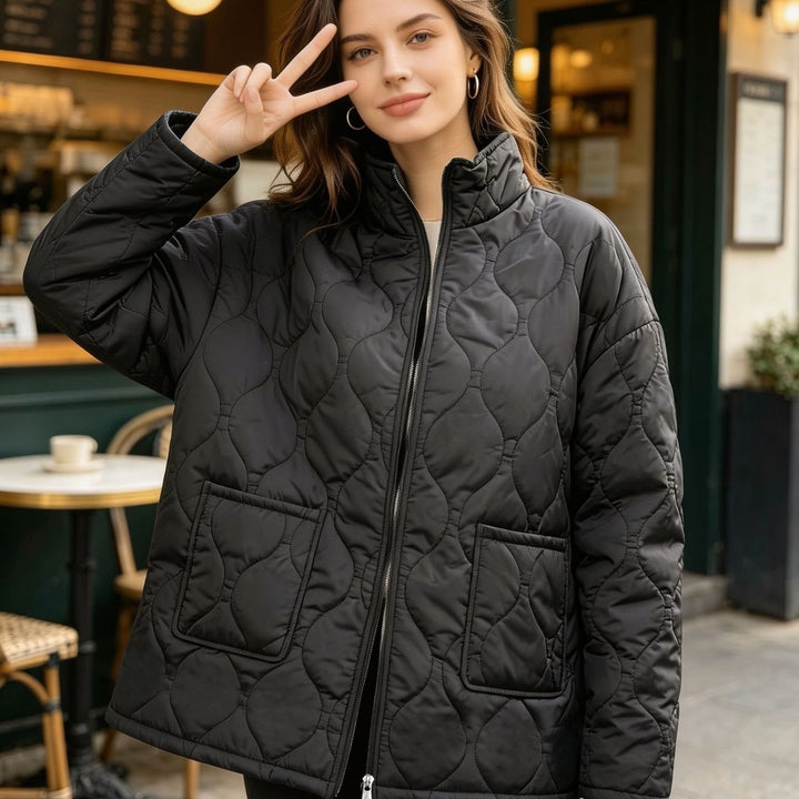 Plus size quilted coat in black shown from the front outside a cafe with a stand collar, zipper and two square patch pockets.