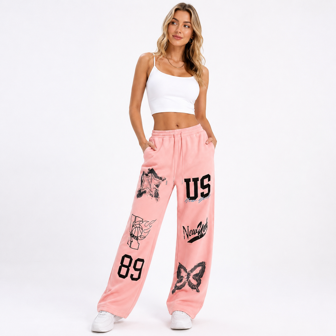 A woman modeling a pair of pink sweatpants with black graphic prints, a white crop top, and white sneakers against a plain white studio background.