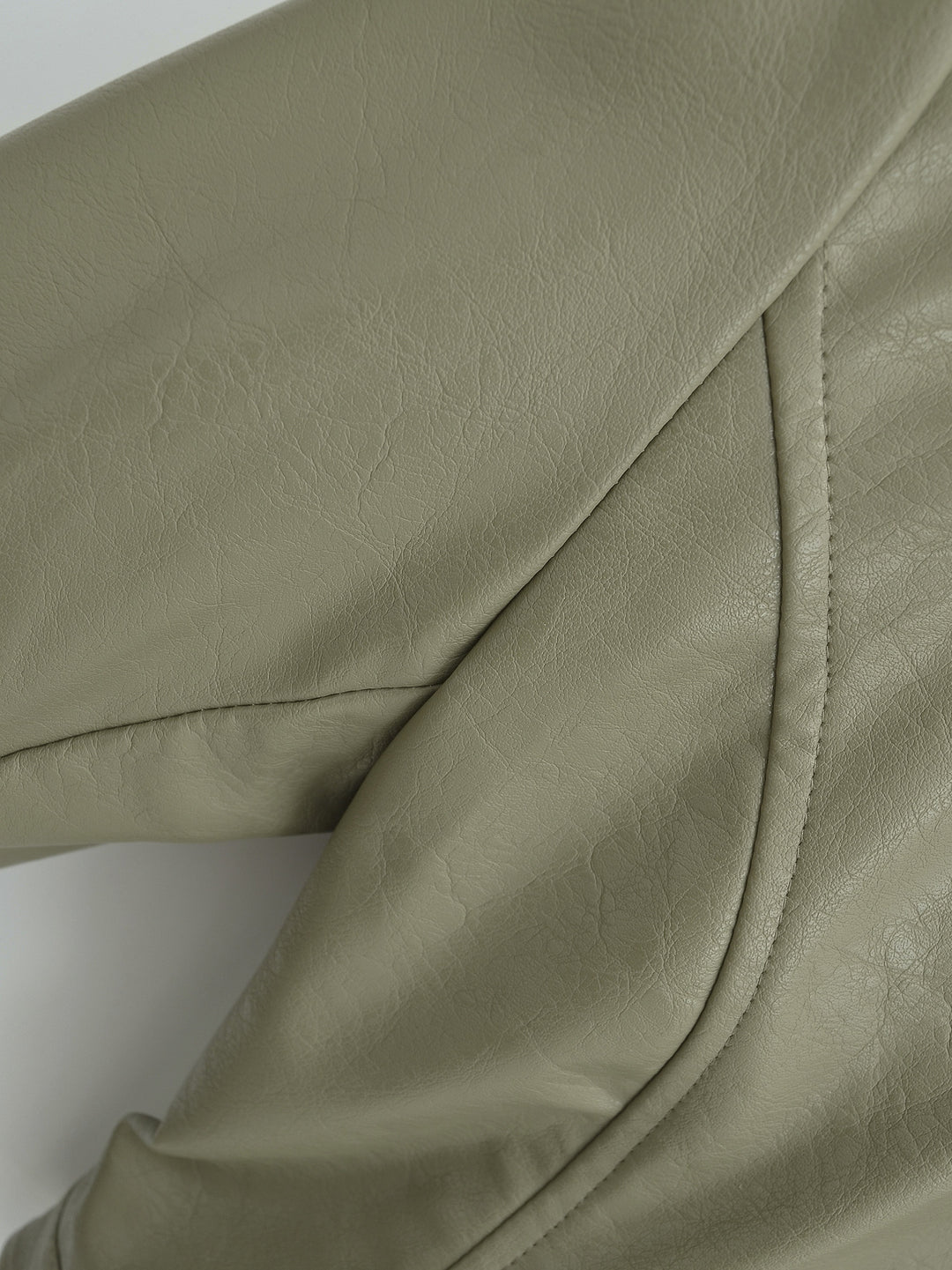 Gray Green jacket shoulder close-up showing faux leather grain, with a curved armhole seam and panel stitching against white.