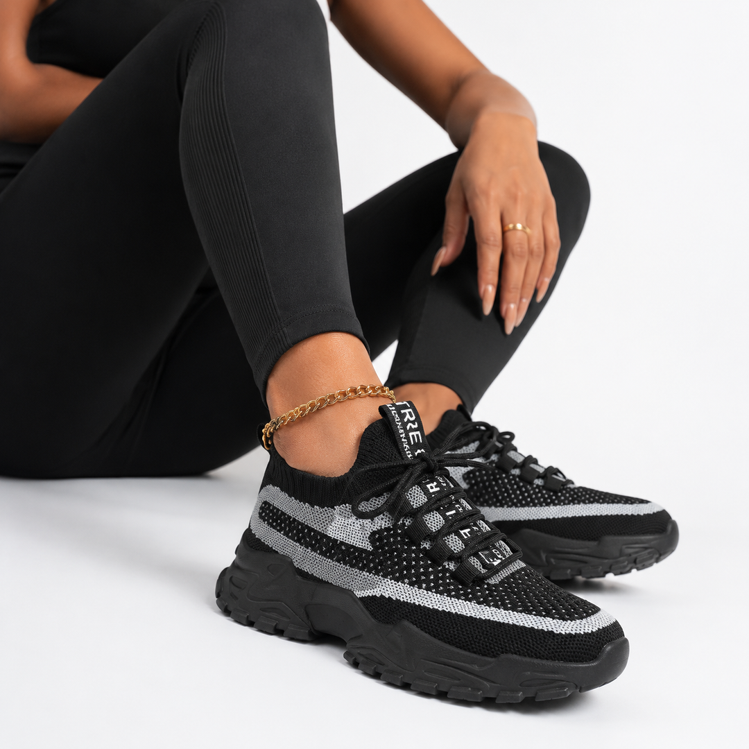A person wearing black ribbed leggings and black chunky sneakers with a silver reflective stripe detail, seated against a clean white studio background.