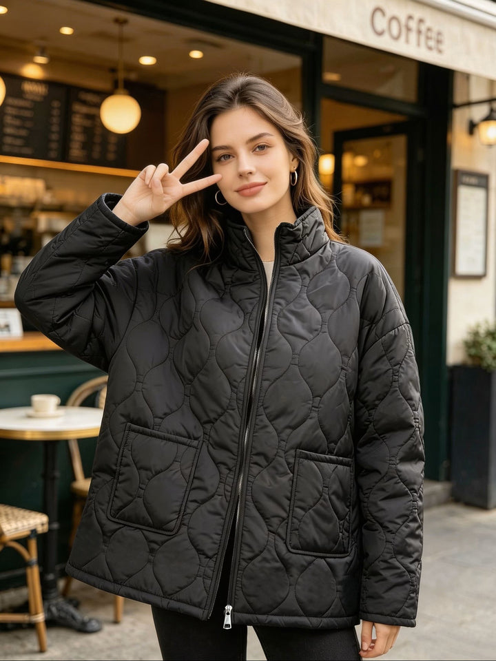 Front detail of the black quilted coat outside a cafe, showing the stand collar, front zip, wavy stitching and patch pockets.