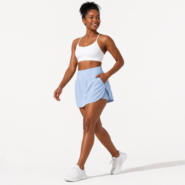 A woman modeling a white sports bra and a light blue athletic skort with white sneakers, posed against a clean white studio background.
