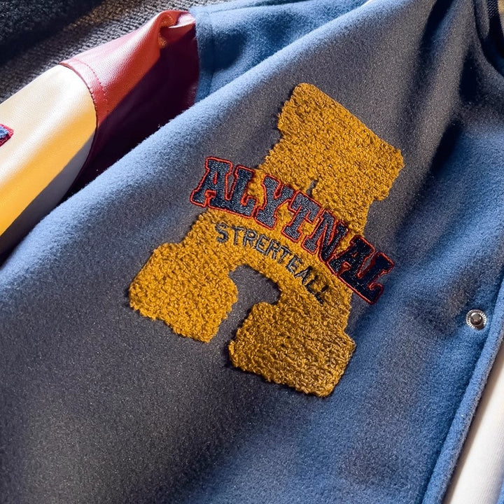 WT167 jacket in Blue shown in a close detail view with a yellow chest patch and embroidery visible, on a gray fabric surface.