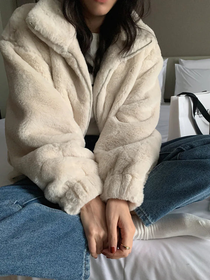 Beige plush jacket in a seated close view with blue jeans, showing the plush texture, a stand collar and full-length sleeves.