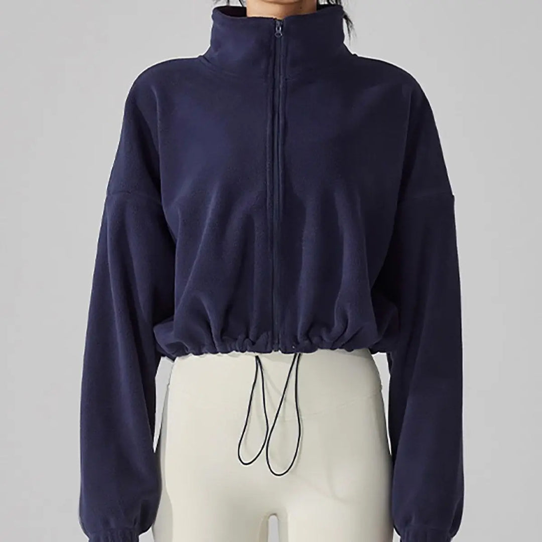 A model wearing a navy blue cropped jacket with a high collar, styled with ivory trousers in front of a gray studio backdrop.