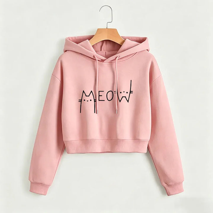 Dusty pink cropped Meow hoodie on a wooden hanger against a light wall, showing a black cat graphic, hood and ribbed sleeves.