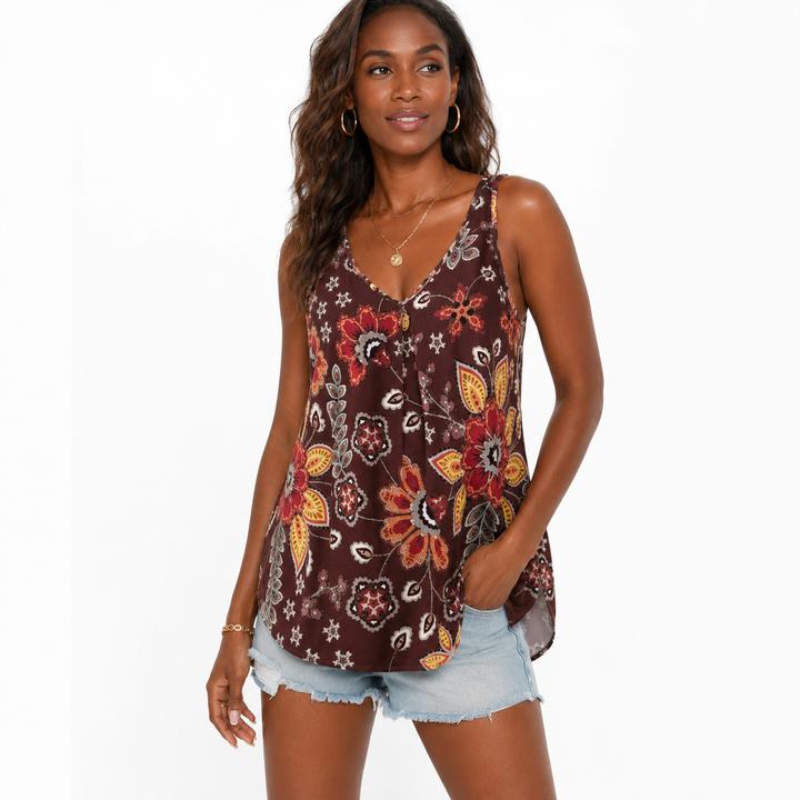 A woman modeling a sleeveless V-neck top with a brown base and a colorful floral and paisley print in red, orange, yellow, and white, paired with denim shorts against a plain white background.