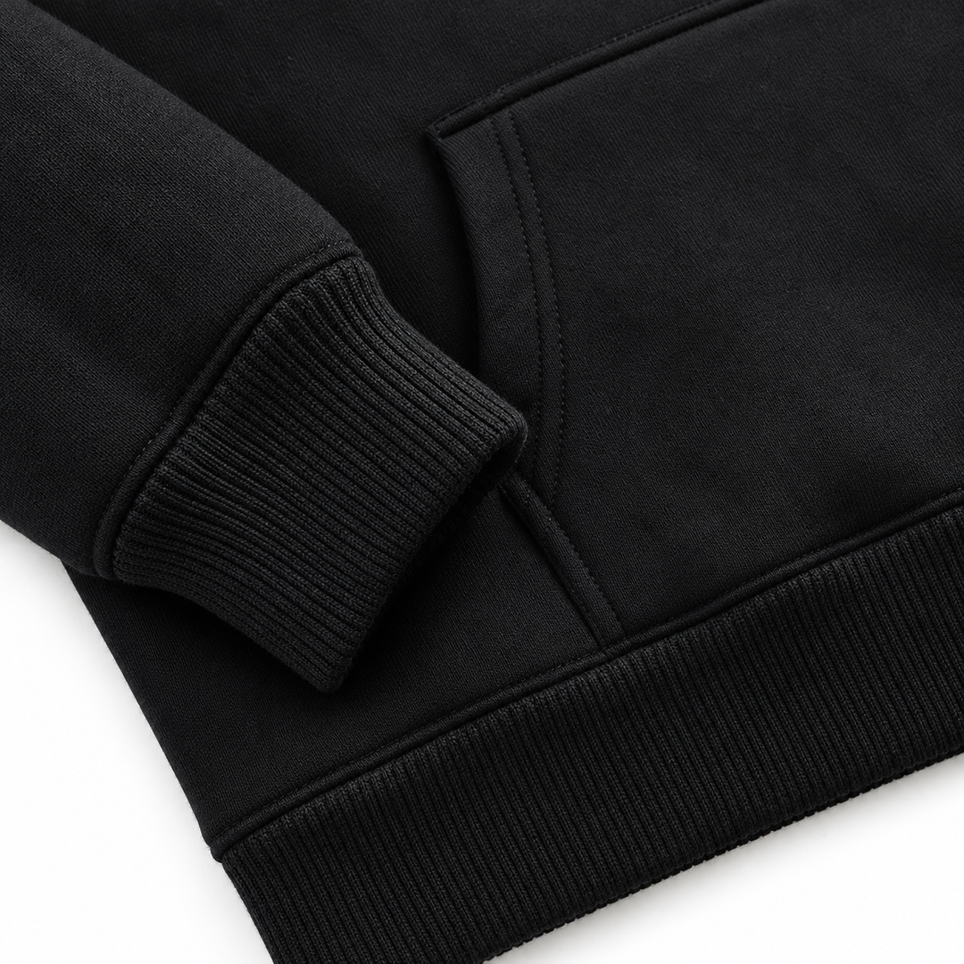 Black pullover hoodie detail showing the ribbed sleeve cuff, kangaroo pocket edge and hem stitching in a bright white studio.