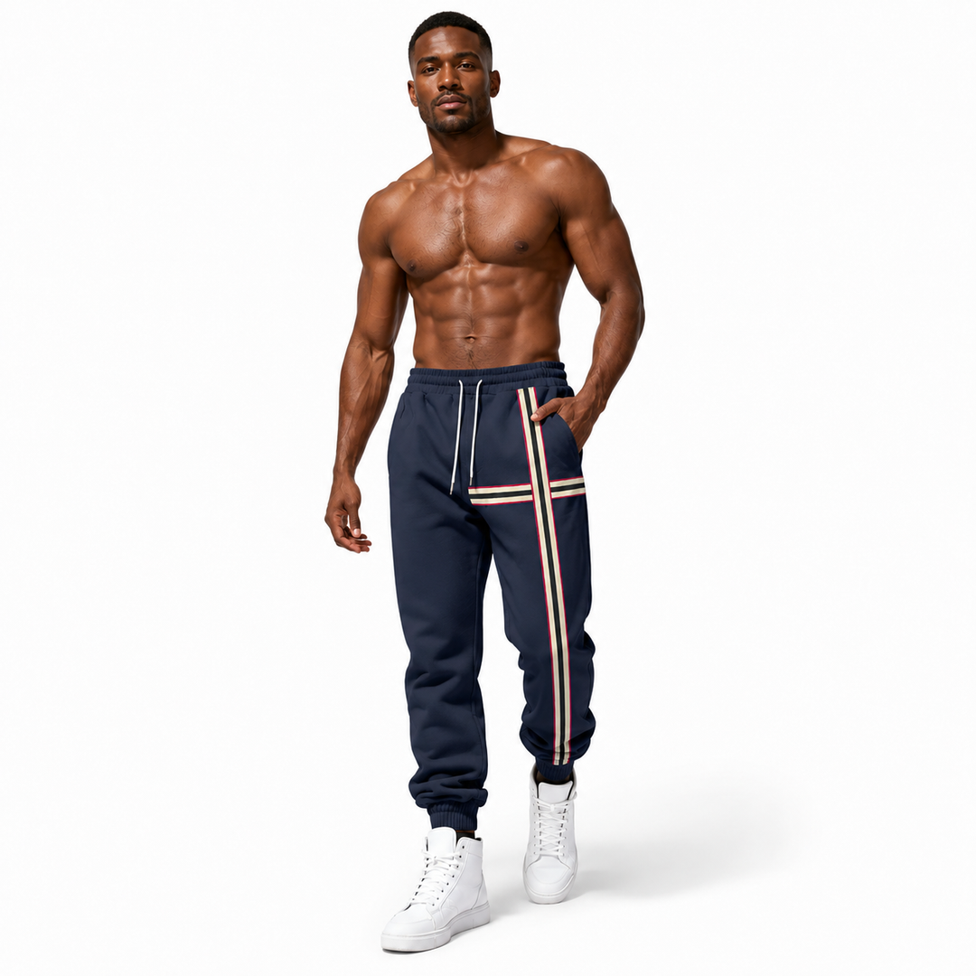 A shirtless muscular man modeling navy blue sweatpants with white and red side stripes, paired with white sneakers, against a plain white background.