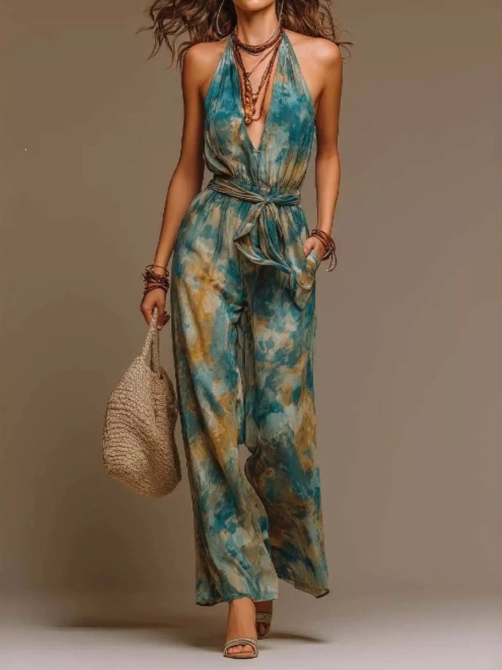 A woman modeling a sleeveless halter-neck jumpsuit with a blue, teal, and beige watercolor-style print. She is holding a woven straw bag and wearing layered necklaces and bracelets, set against a plain beige studio background.