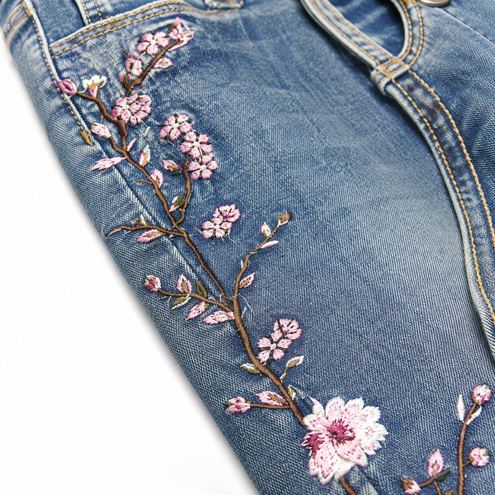 Close-up of pink floral embroidery on blue denim, showing thread details, branch stitching and textured denim weave on white.
