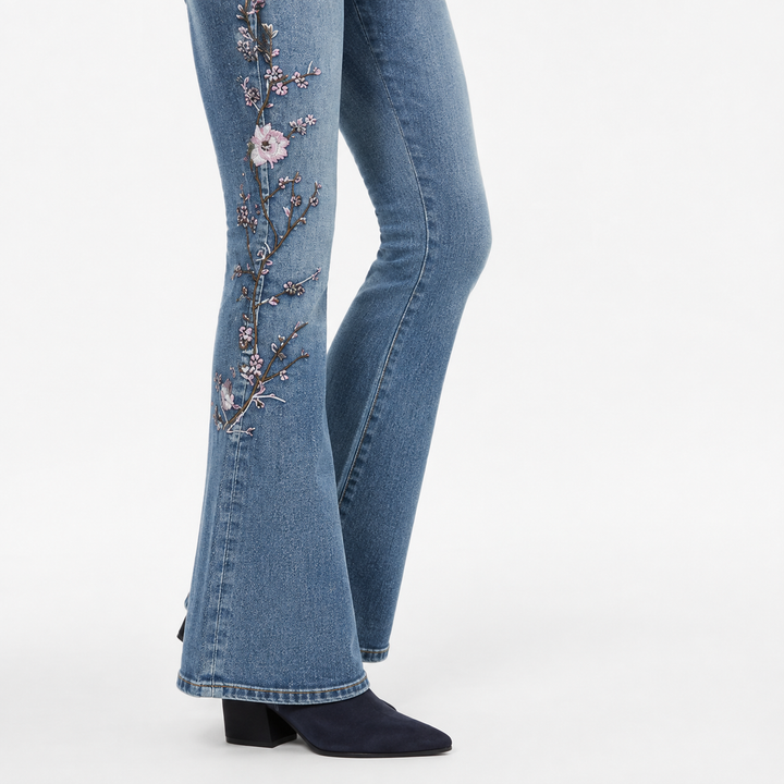 Lower-leg view of embroidered flared jeans fit showing floral branch, wide hem finish and blue denim texture over dark boots.