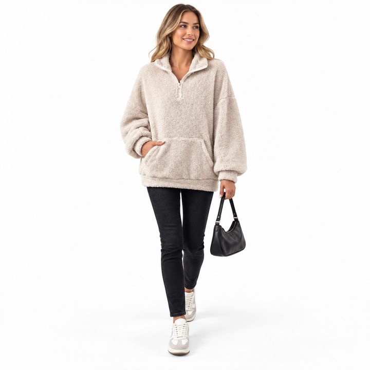 Beige fleece pullover styled with jeans, trainers and a black handbag on a full-length model taking a step in a white studio.