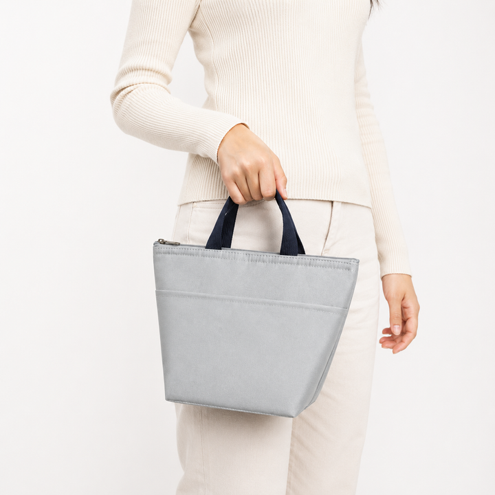 Light gray portable lunch tote carried by a woman, showing compact proportions, short navy handles and simple everyday shape.