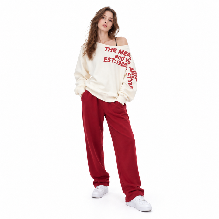 Apricot pullover with red lettering on a full-body model wearing red trousers and white trainers, shown on a studio backdrop.