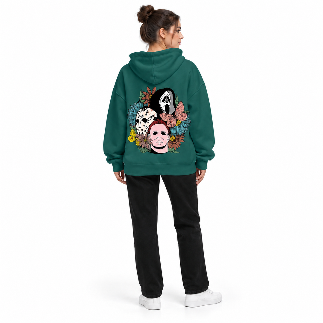 Full-body rear view of a woman wearing the dark green hoodie with a mask and flower print, black trousers and white sneakers.