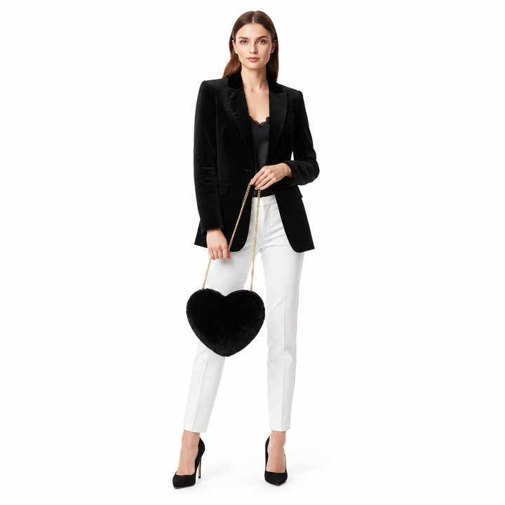 Full-body woman in a black blazer and white trousers holding a black plush heart bag with a gold-tone chain in a white studio