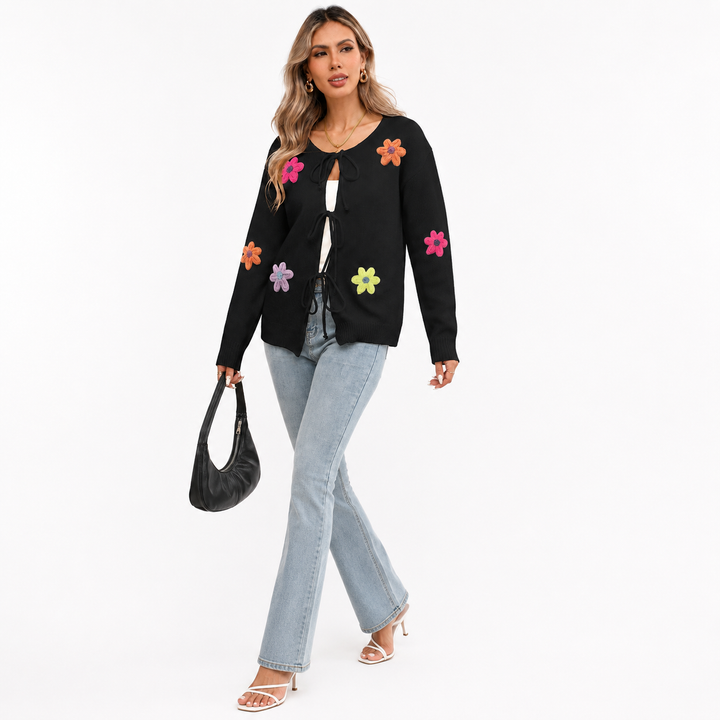Black floral tie-front knit cardigan on a woman in a walking studio pose with colorful flower appliques and light-wash jeans.