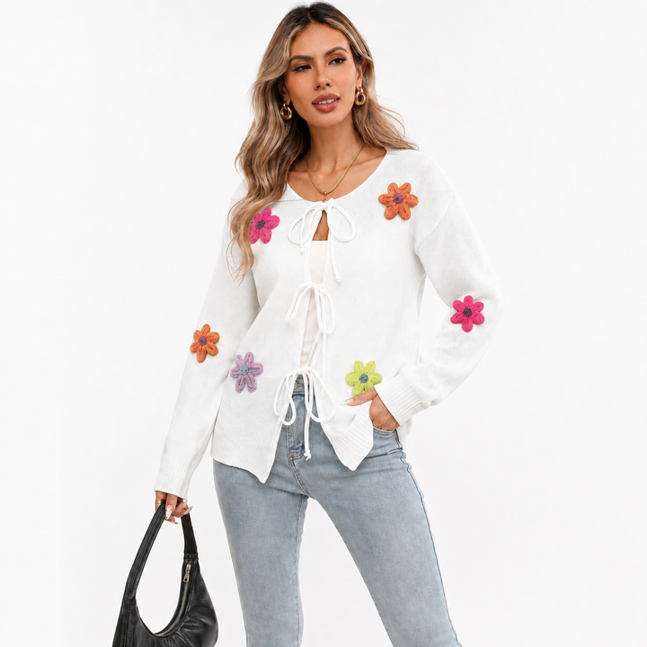 White floral tie-front knit cardigan on a woman with visible face, light-wash jeans, colorful flower appliques and black handbag.