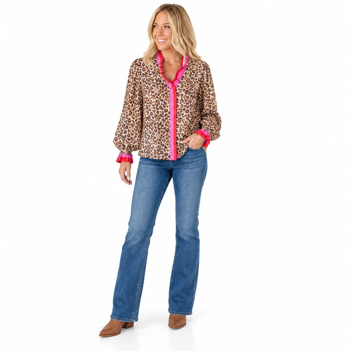 Soft Pink Leopard Glow blouse with pink and red ruffled trim, worn with blue jeans and brown shoes in a full body studio view