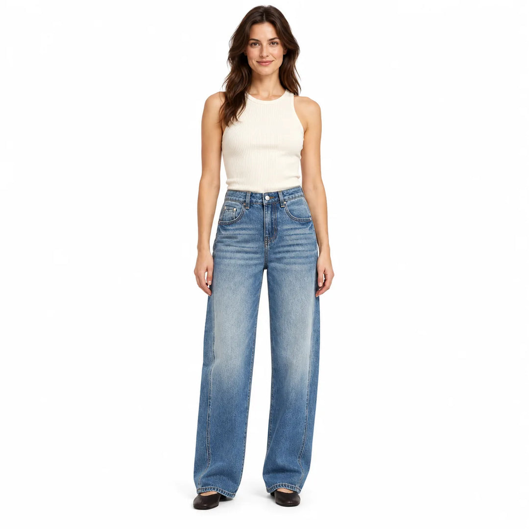 Brunette woman wearing blue washed wide leg jeans with a cream sleeveless top and dark flats, shown against a white backdrop.