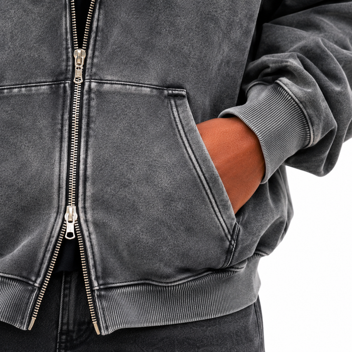 Detailed view of washed charcoal hoodie showing metal front zipper, divided pocket opening, ribbed cuff and ribbed lower hem.