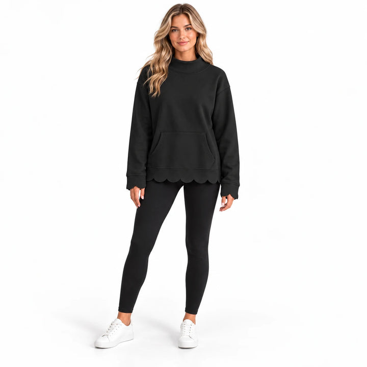 Studio model wearing a black mock neck sweatshirt with scalloped cuffs and hem, styled with black leggings and white sneakers