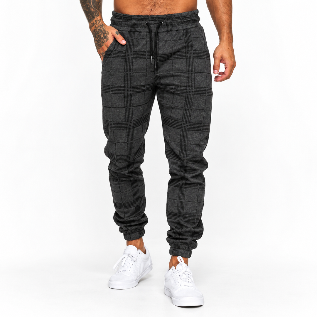 Plaid pants for men in dark grey on male model, front studio view with tapered jogger shape, black drawstring and white shoes