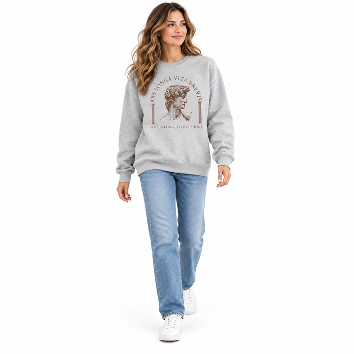Gray art print sweatshirt on a woman walking in a white studio shown head to shoes with blue jeans and white casual footwear.