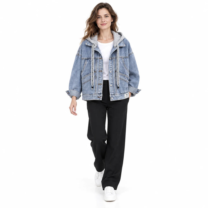 Adult model walking in a white studio wearing a blue denim jacket with grey hood, white top, black trousers, and white shoes.