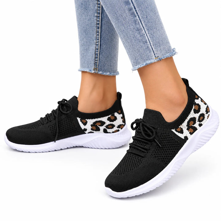 Black mesh lace-up sneakers worn with cropped blue jeans show white soles and leopard heel panels on this white studio floor.