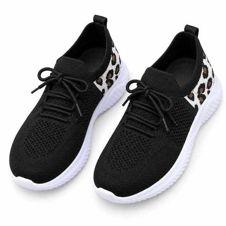 Top view of black lace-up mesh sneakers with round toes, white soles and patterned panels, placed together in a white studio.
