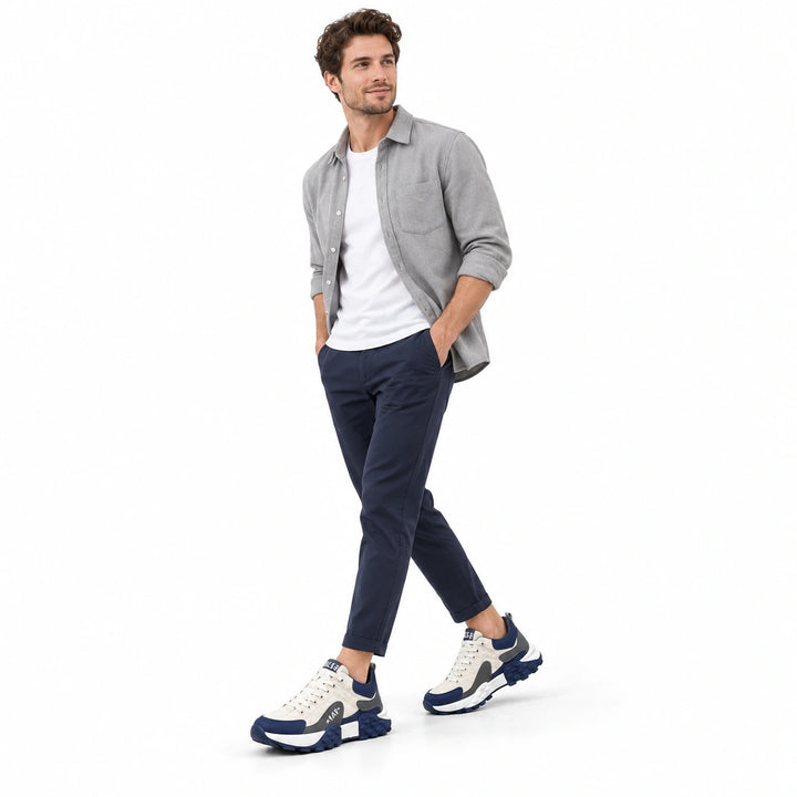 Full-body male model wearing White and Navy sneakers with a gray overshirt, white T-shirt and navy trousers on a white stage.