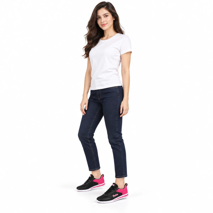 Full-body woman wearing black and pink mesh sneakers with a white T-shirt and dark jeans, standing in a bright, white studio.