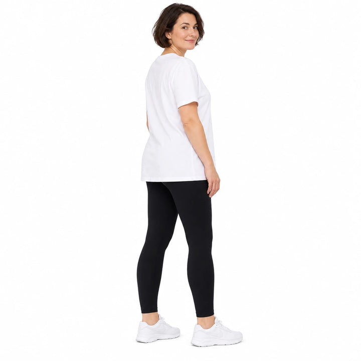 Short-haired adult model in black leggings and white longline T-shirt, standing with white trainers on white studio backdrop.