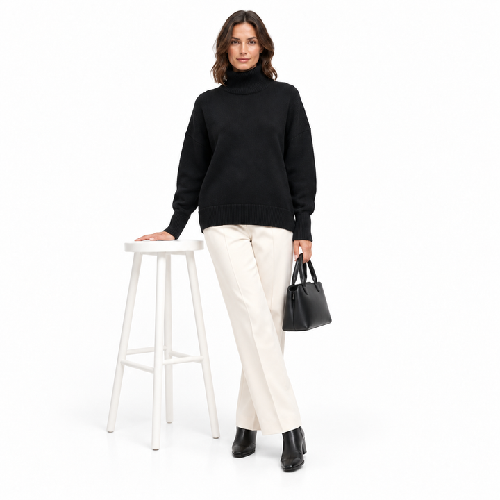 Studio outfit with a model wearing the black turtleneck sweater beside a white stool, carrying a small plain handbag on white