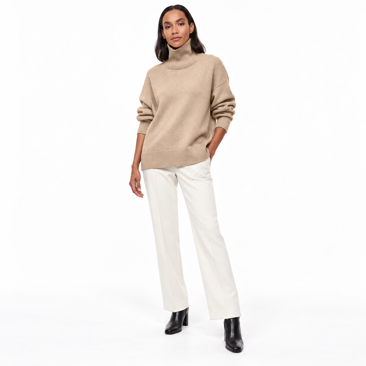 Full-length model wearing a light khaki ribbed-trim turtleneck sweater with ivory tailored trousers and black boots on white.