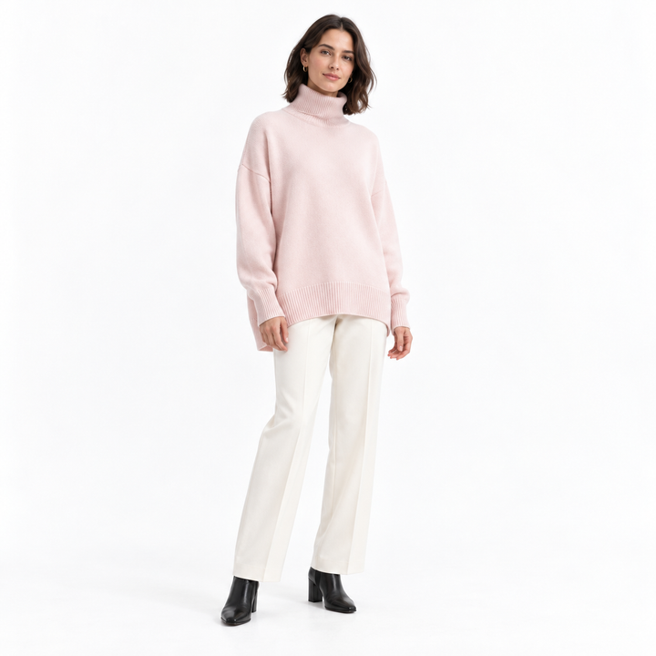 Full-length model wearing a pale pink turtleneck sweater with ivory trousers and black ankle boots in a white studio setting.