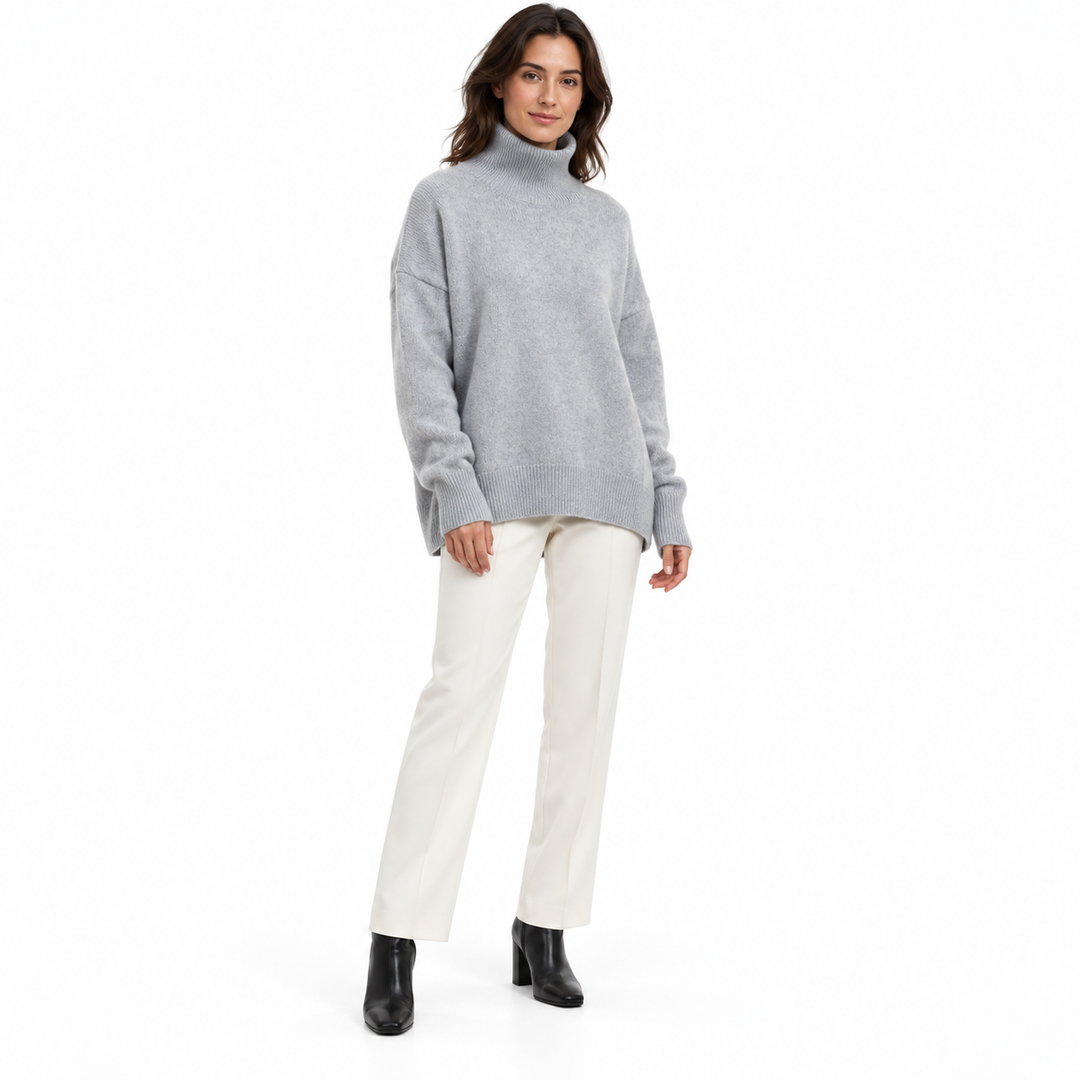 Full-length model wearing a silver gray ribbed-trim turtleneck sweater with ivory tailored trousers and black boots on white.