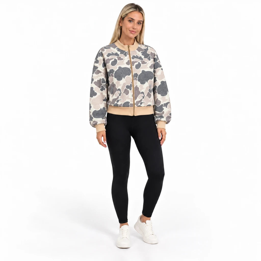 Full-body front view of a blonde model wearing the camouflage bomber with black leggings and white trainers in a white studio