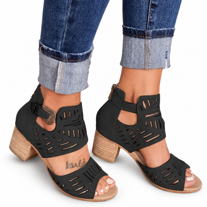 Black block heel sandals with cutout panels and side buckles, worn with cuffed jeans on a white background with soft shadows.