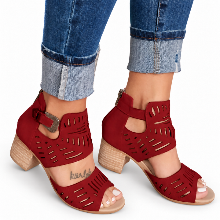 Red cutout sandals with side buckles, open toes and separate broad heels, shown with cuffed jeans against a white background.
