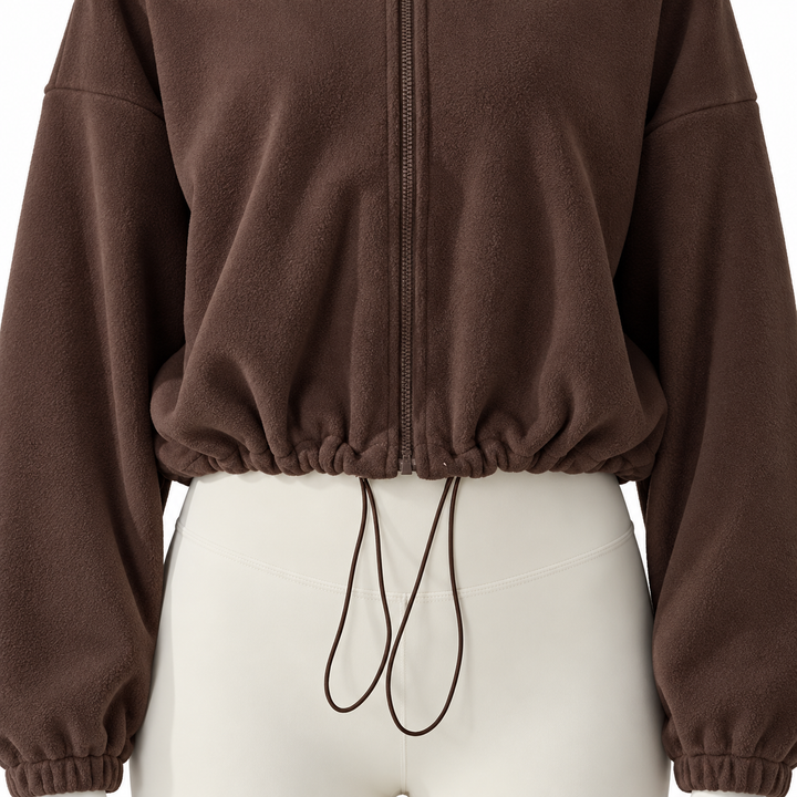 Dark brown jacket detail showing the lower front zipper, gathered cropped hem and long drawcord loops against ivory trousers.
